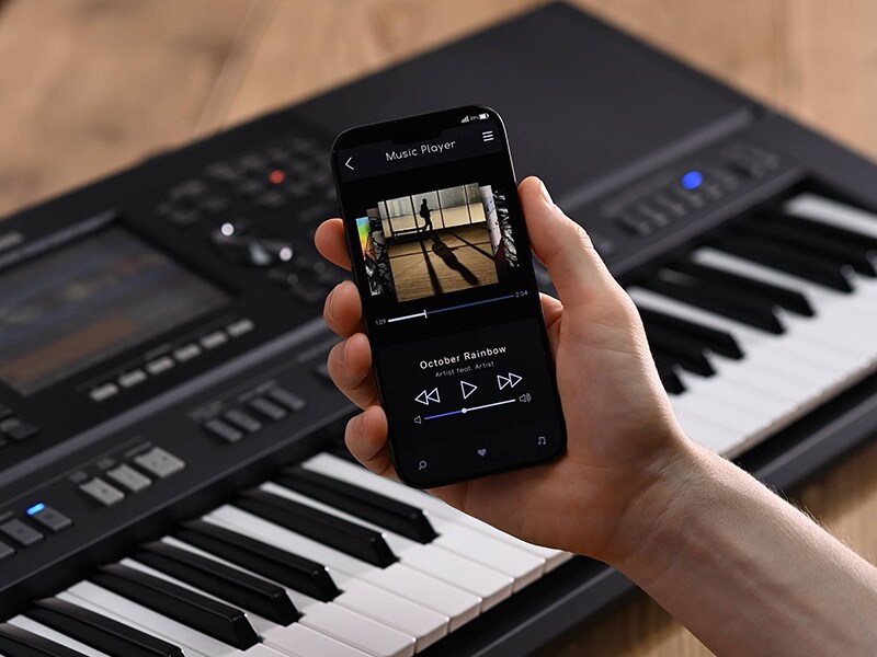 Yamaha Arranger Workstation PSR-SX920 dan tangan yang memegang smartphone dengan tampilan layar pemutar musik