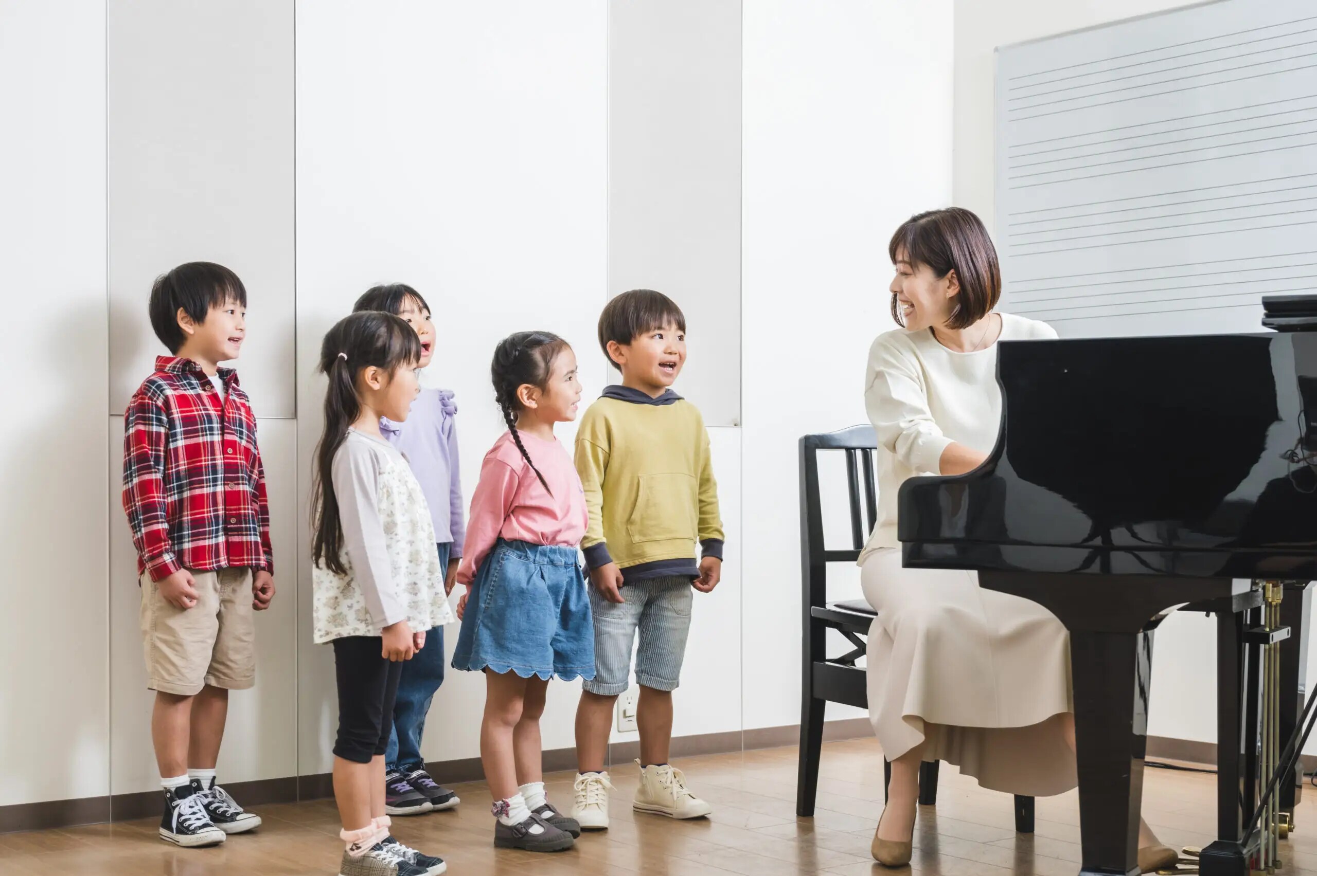A group of young children standing and singing near a grand piano, while a smiling female music teacher plays the piano and interacts
                                with them in a bright music classroom with a whiteboard in the background.