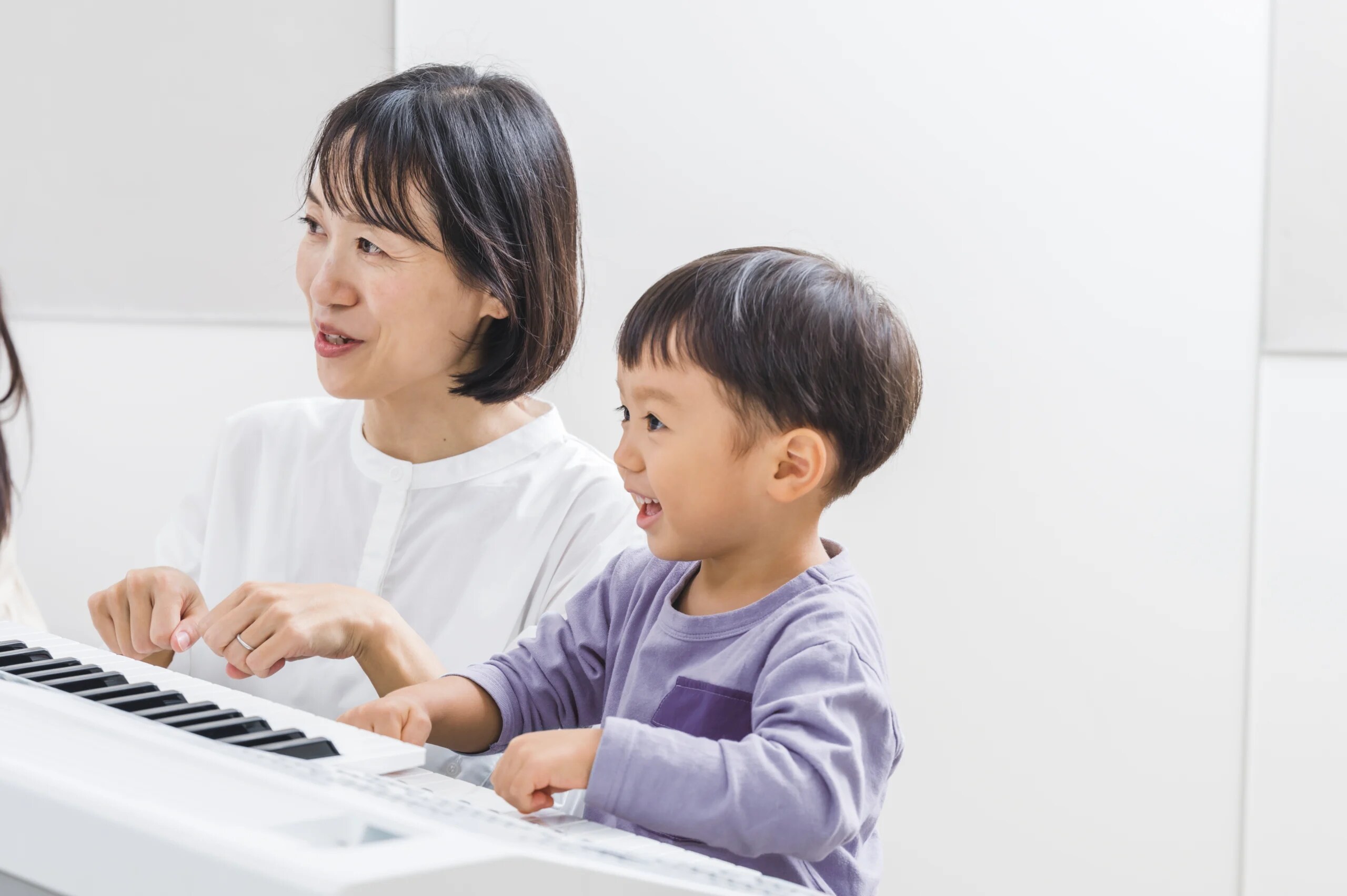 A smiling young child learning to play keyboard with a supportive female teacher at Yamaha Music School – joyful early music education
                                for children.