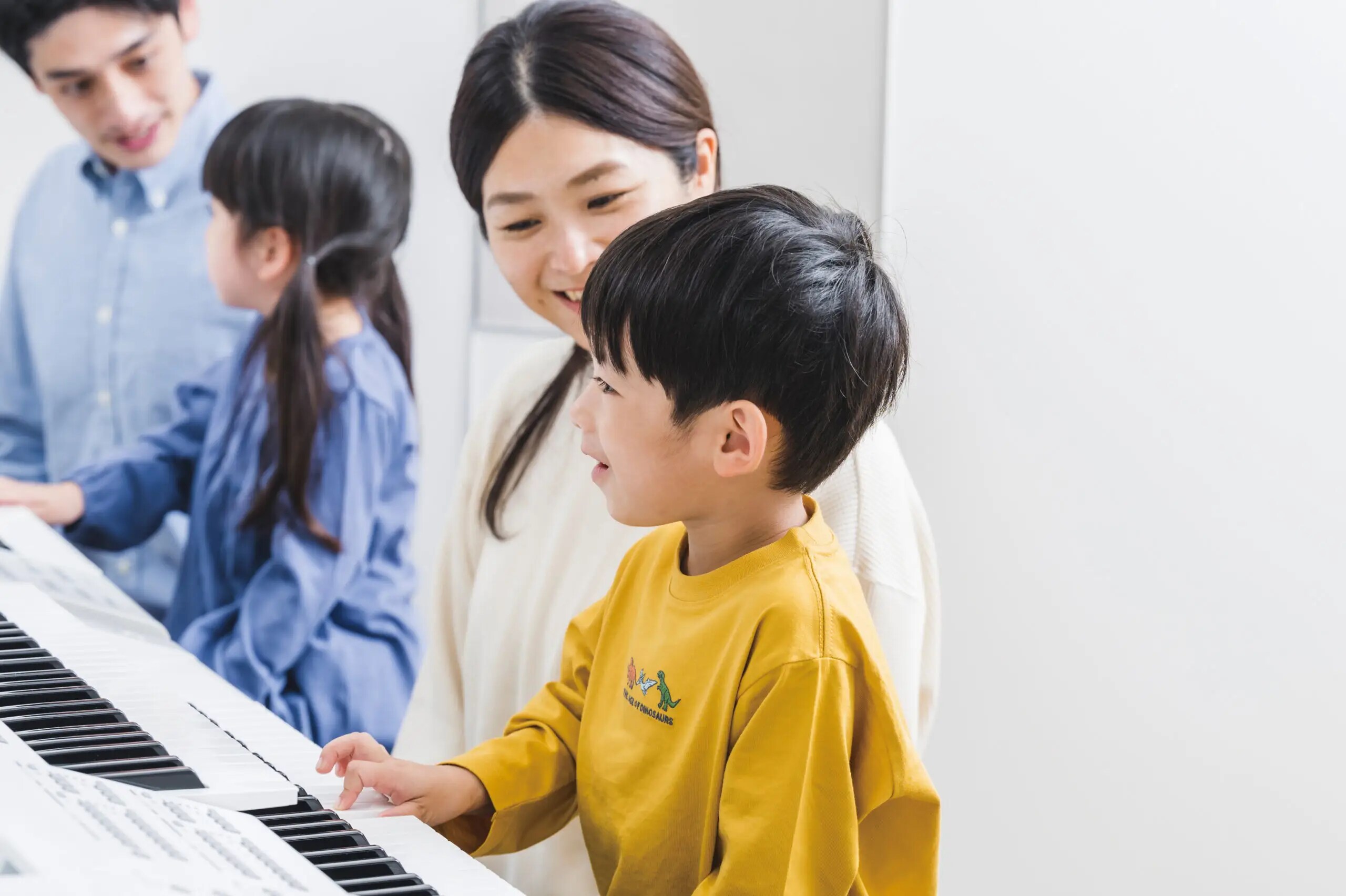 Young child in yellow jumper playing piano with mother sitting beside him, while another child plays electone
                                with father in the background at Yamaha music school