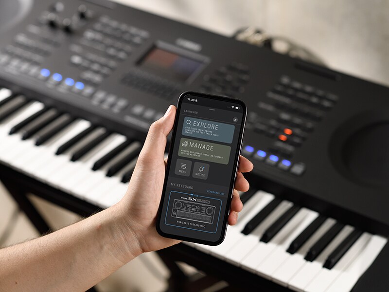 PSR-SX920 dan tangan yang memegang smartphone dengan layar pemutar musik.